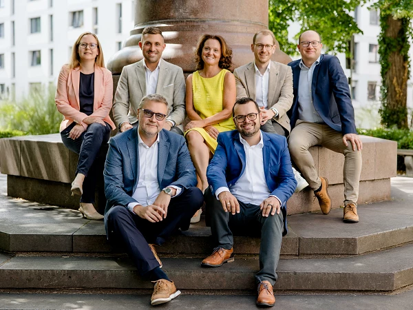 Eine Gruppe von sieben Personen sitzt und steht auf einer Treppe in einem begrünten, urbanen Umfeld. Sie lächeln in die Kamera, während sie in formeller Kleidung gekleidet sind. In der Mitte sitzt ein Mann in einem blauen Blazer, umgeben von anderen Personen in hellen und neutralen Tönen. Im Hintergrund sind moderne Gebäude und Bäume sichtbar.