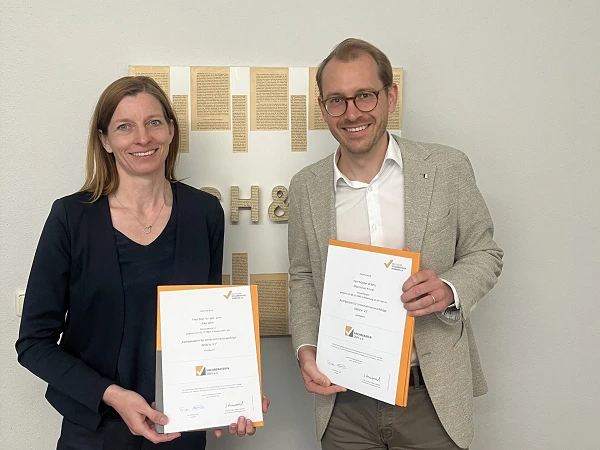 Zwei Personen stehen nebeneinander vor einem Hintergrund mit einem goldenen Logo. Beide halten je ein Zertifikat in der Hand und lächeln in die Kamera.