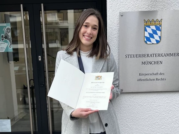 Eine junge Frau hält stolz ein Zertifikat in der Hand und steht vor der Eingangstür der Steuerberaterkammer München, die mit einem Schild und dem bayerischen Wappen versehen ist.