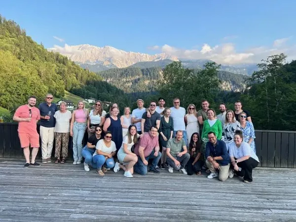 Eine große Gruppe von Menschen steht auf einer Holzterrasse vor einer malerischen Berglandschaft mit grünen Bäumen und einem klaren Himmel. Die Gruppenmitglieder lächeln und posieren zusammen für das Foto.
