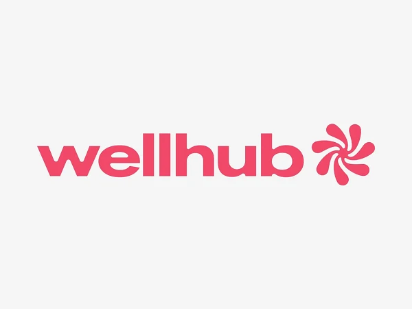 Das Bild zeigt das Logo von "wellhub", das aus dem Schriftzug "wellhub" in pinker Schriftfarbe und einem stilisierten blütenförmigen Symbol auf der rechten Seite besteht. Hintergrund ist weiß.