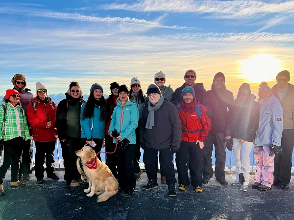 Eine Gruppe von 17 Personen steht auf einer Aussichtsplattform im Schnee, umgeben von Bergen, während die Sonne im Hintergrund aufgeht. Ein Hund mit einem roten Halstuch sitzt vor der Gruppe. Die Menschen tragen Winterkleidung und lächeln in die Kamera.