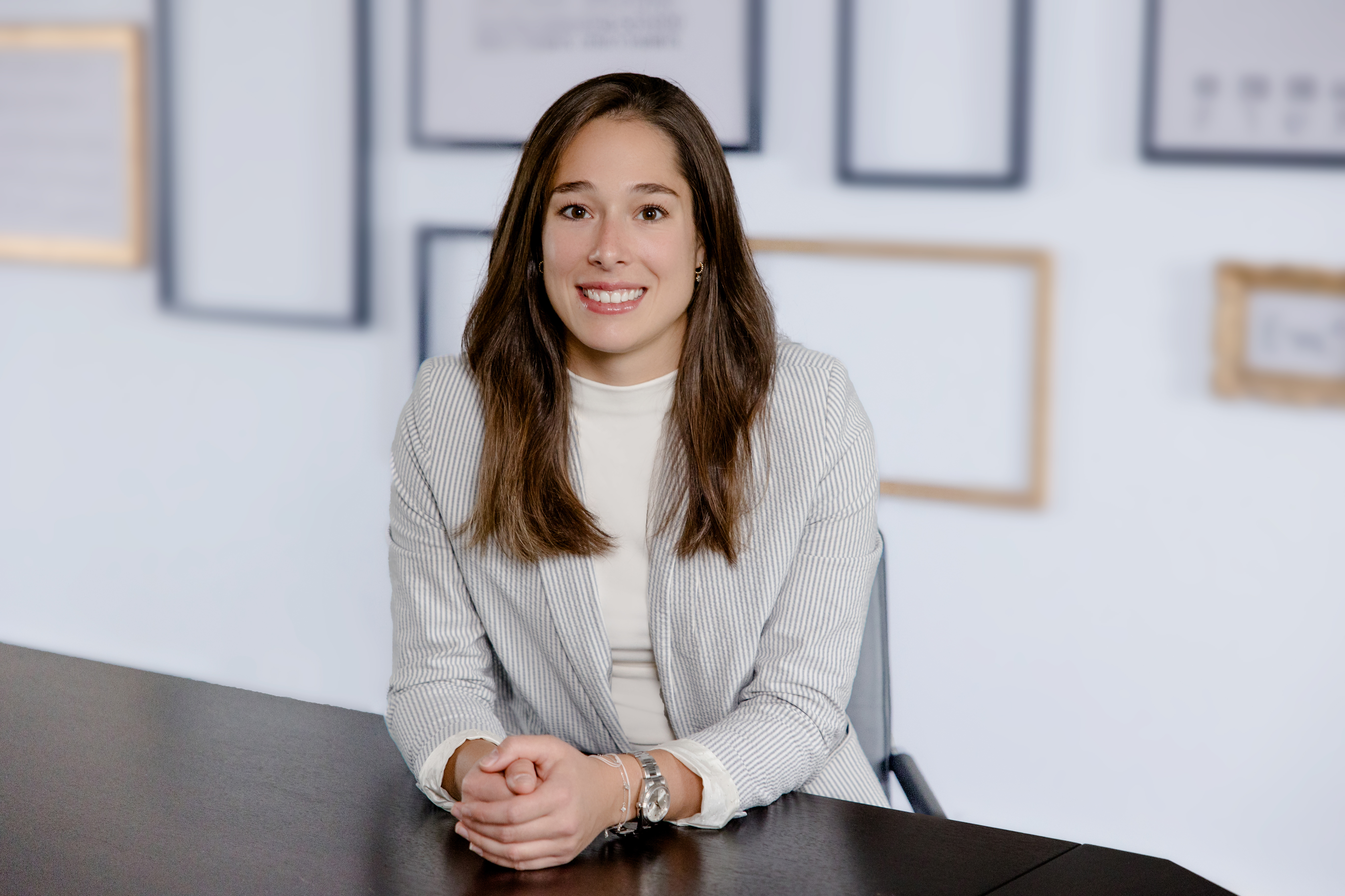 Eine lächelnde Frau mit langen, braunen Haaren sitzt an einem Tisch und trägt einen Streifenanzug, umgeben von einem minimalistischen Hintergrund mit Bilderrahmen.