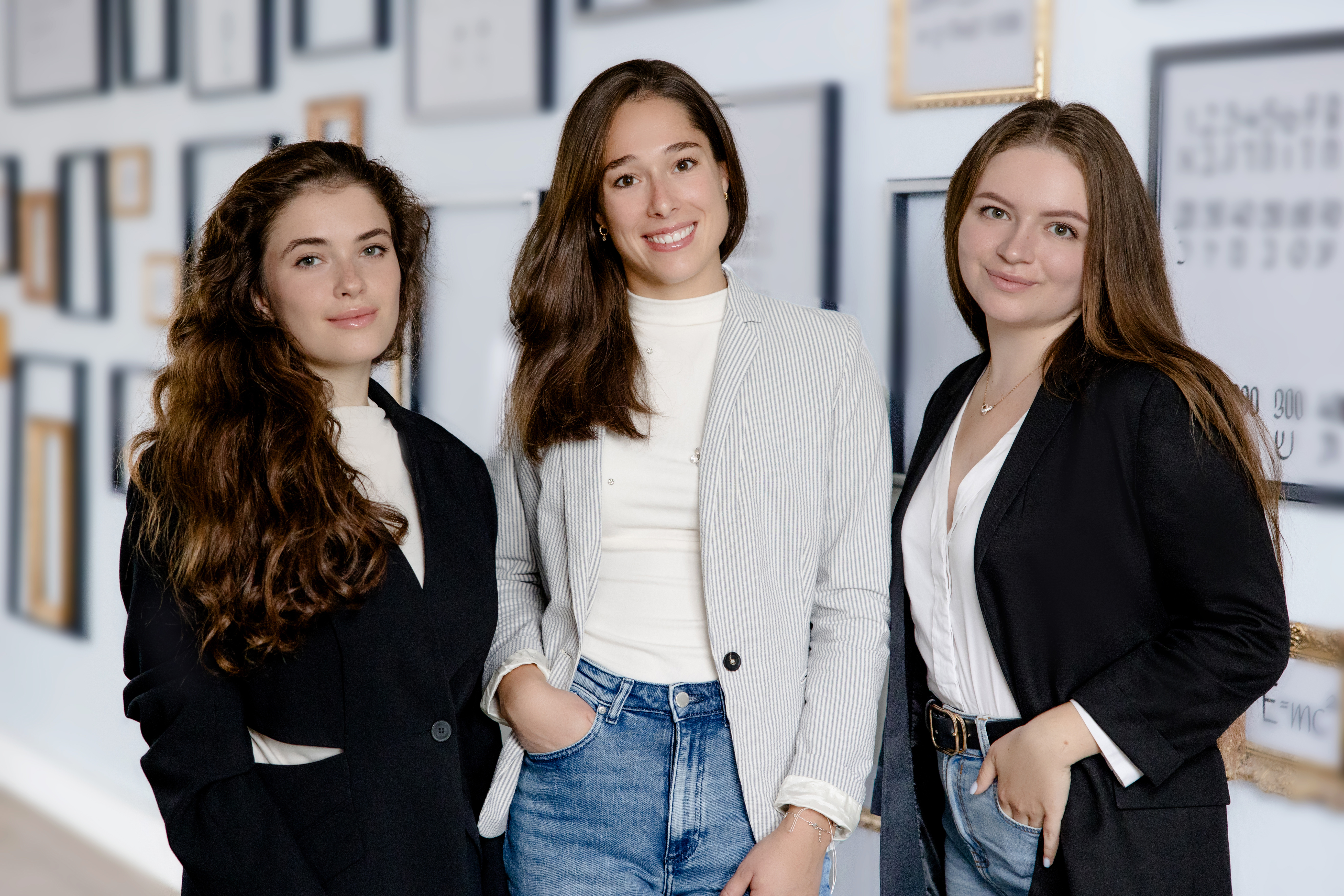 Drei junge Frauen stehen lächelnd in einem modern gestalteten Raum mit gerahmten Bildern im Hintergrund. Die erste Frau hat lange, lockige Haare und trägt einen schwarzen Blazer. Die zweite Frau hat mittellange, glatte Haare und trägt einen grauen Blazer. Die dritte Frau hat braune Haare und trägt ebenfalls einen schwarzen Blazer.