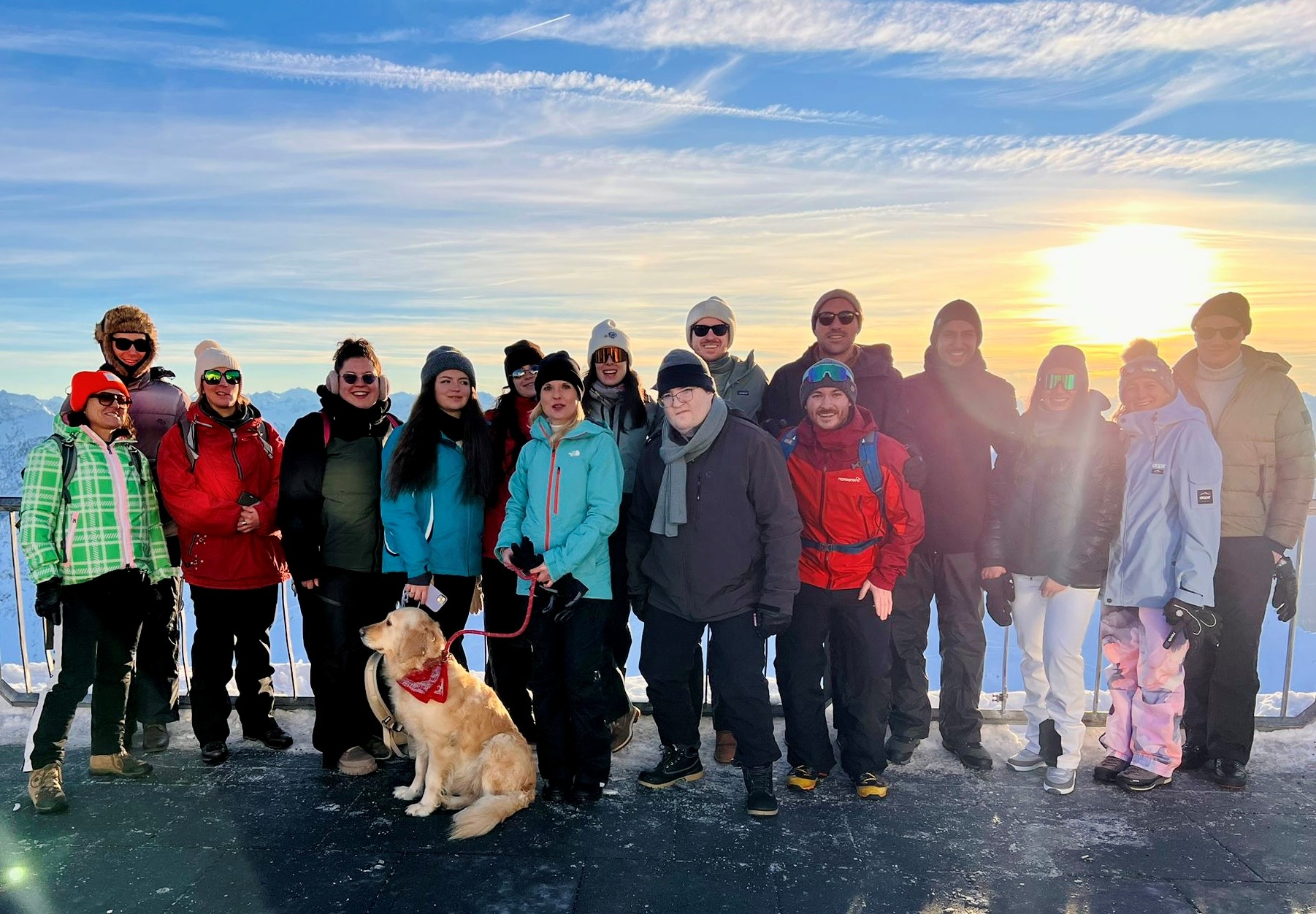 Eine Gruppe von 17 Personen steht auf einer Aussichtsplattform im Schnee, umgeben von Bergen, während die Sonne im Hintergrund aufgeht. Ein Hund mit einem roten Halstuch sitzt vor der Gruppe. Die Menschen tragen Winterkleidung und lächeln in die Kamera.